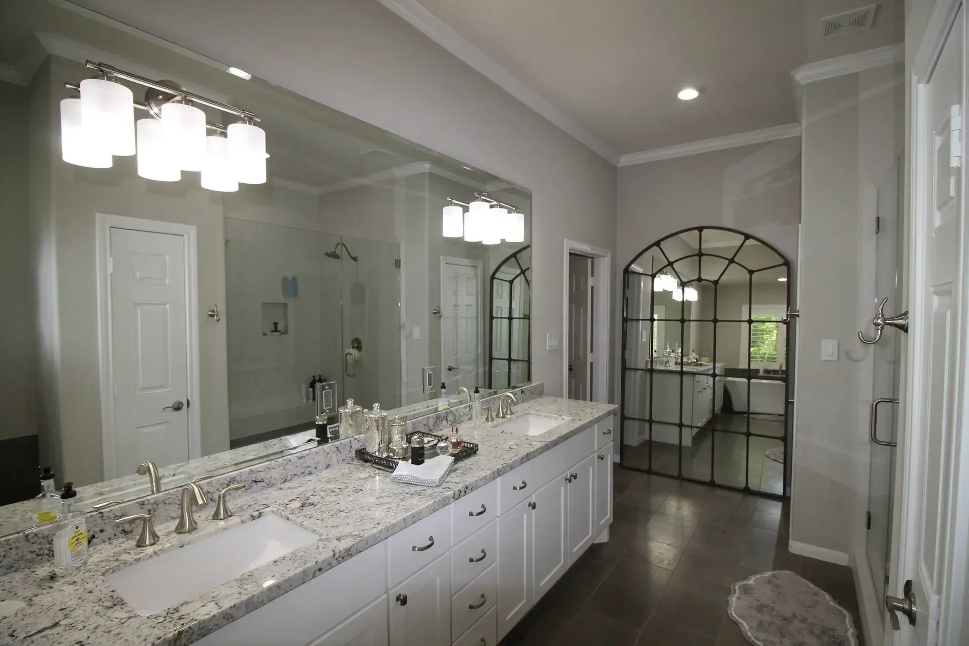 Bright bathroom with double vanity, large mirror, white cabinets, and arched doorway to a lit shower area