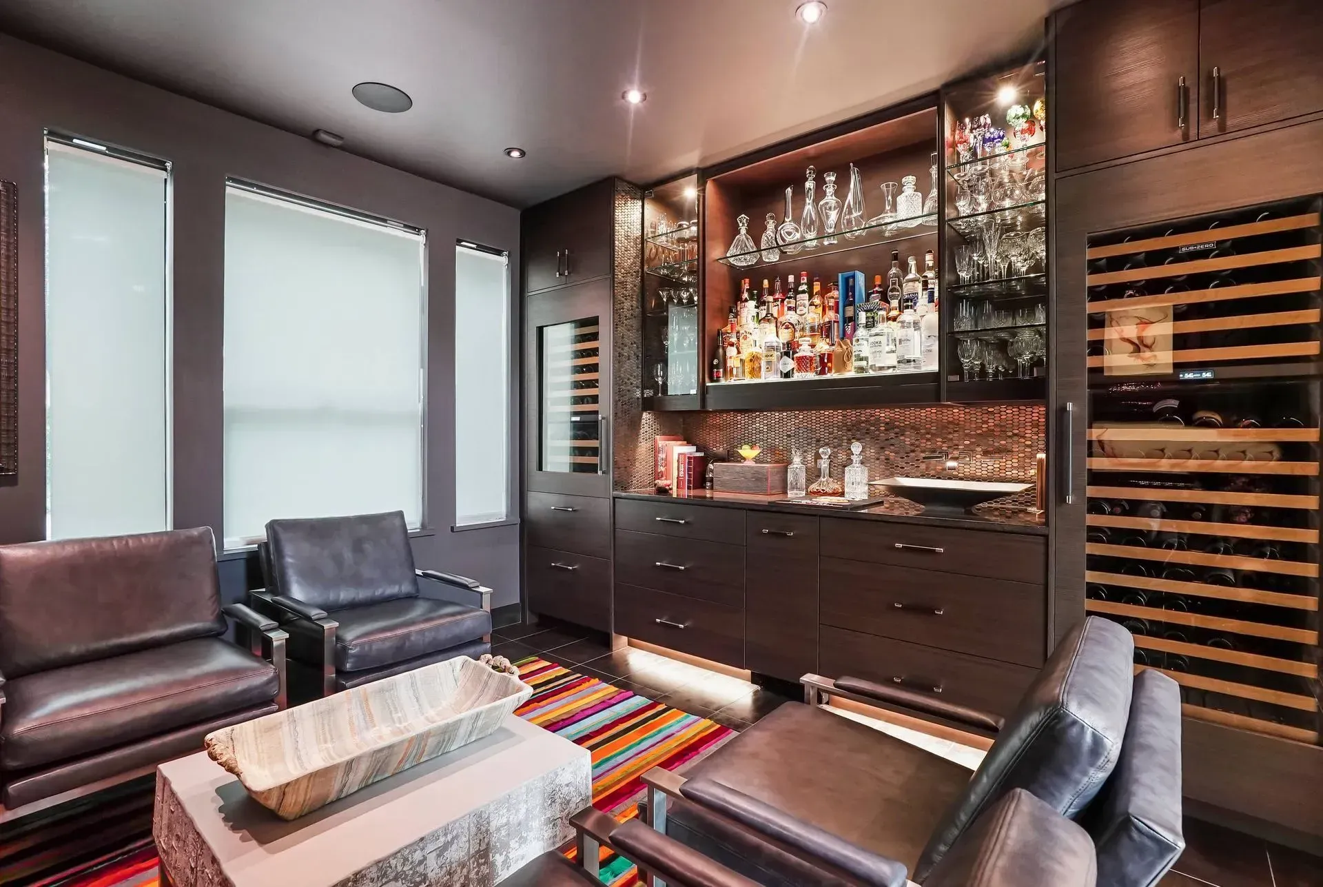 Luxurious home bar lounge with dark wood cabinetry, wine rack, leather chairs, and colorful striped rug