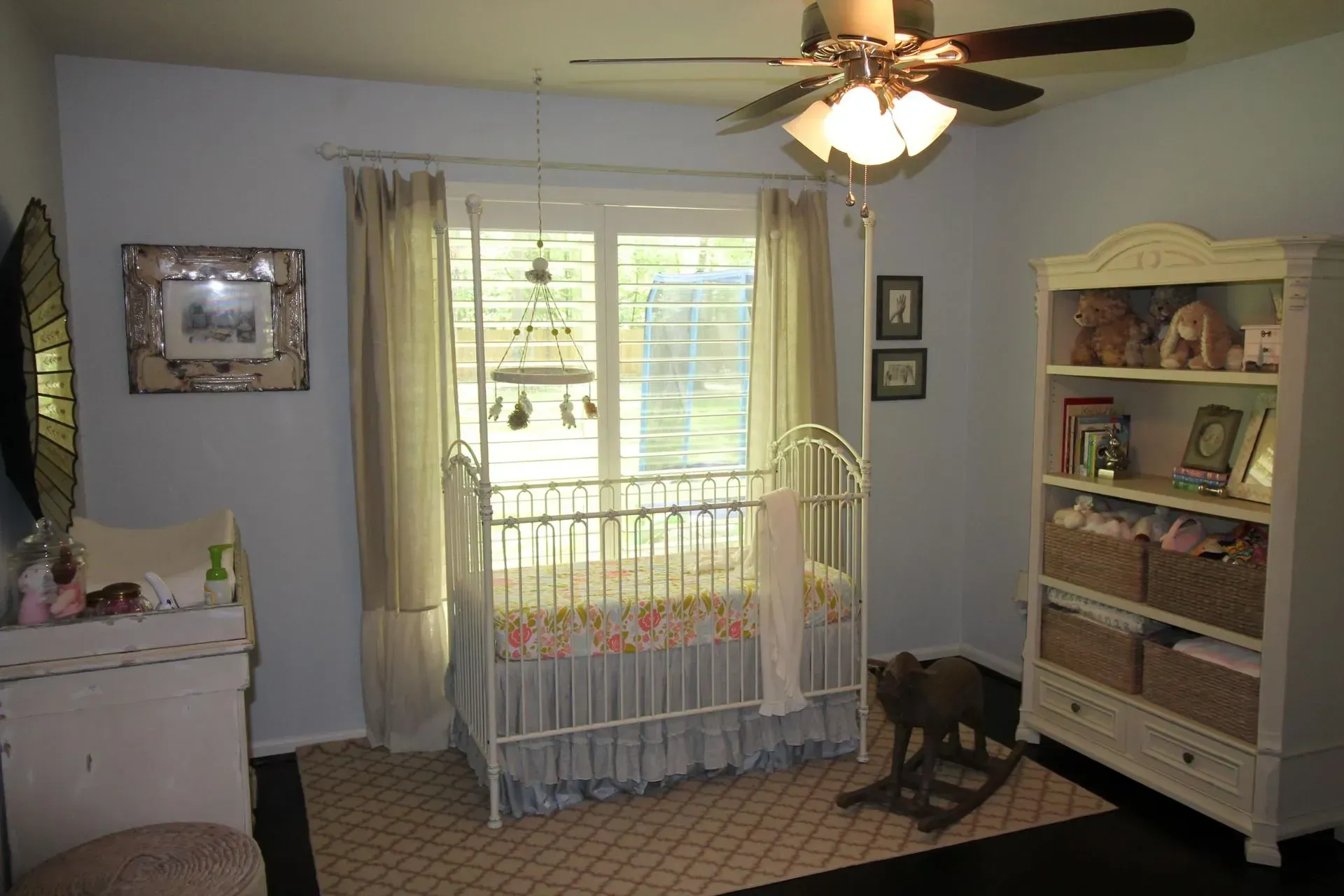Nursery room with crib by window, dresser, and white shelving unit; ceiling fan above and carpeted floor.