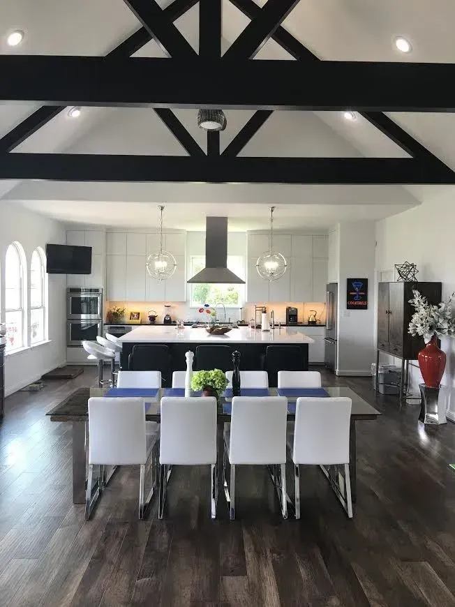 Modern open-plan kitchen and dining area with white chairs, dark wood floors, and exposed black beams.