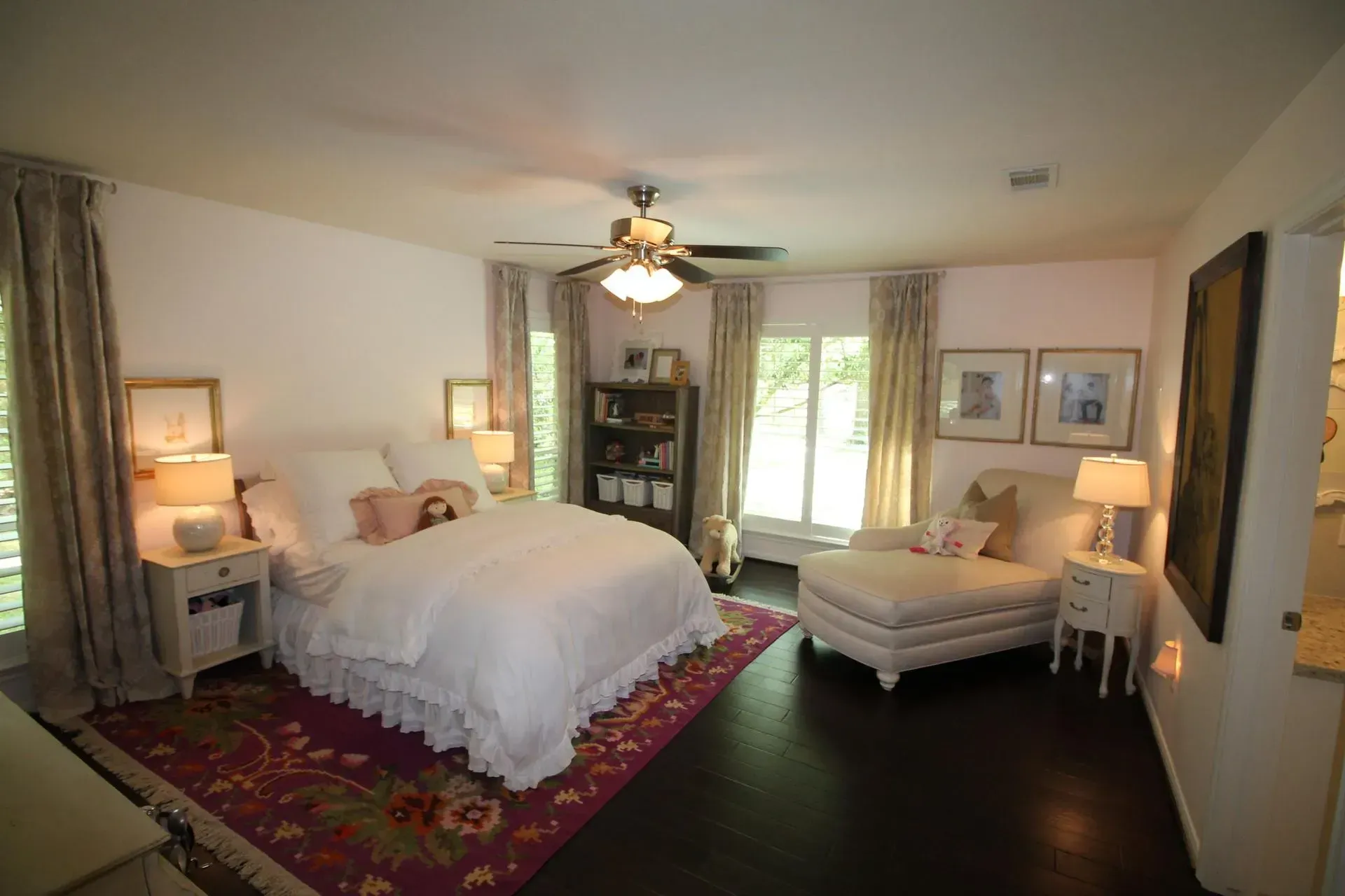 Bright bedroom with white bed, red rug, dark wood floor, and windows overlooking greenery.