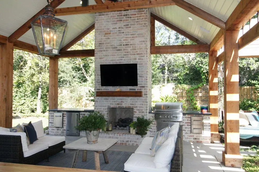 Covered outdoor patio with white sofas, a brick fireplace, and a mounted TV in a wood-beamed pavilion