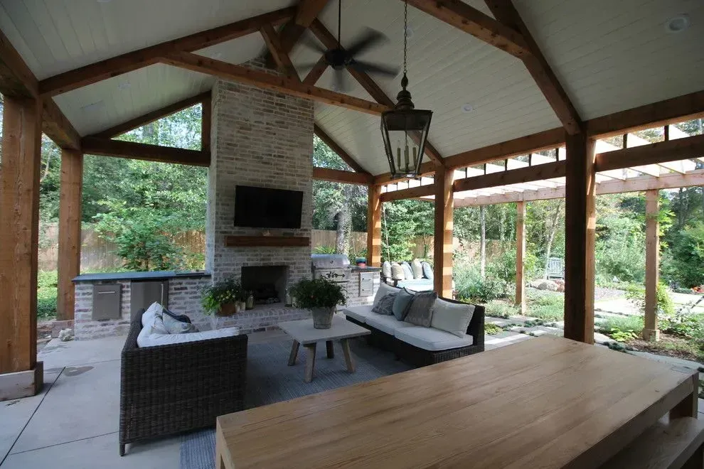 Open-air living room with wicker seating, fireplace, and large wooden table overlooking a lush garden