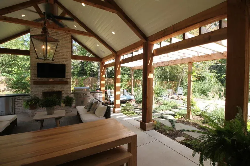 Covered outdoor patio with wooden beams, stone fireplace, cushioned seating, and garden view.