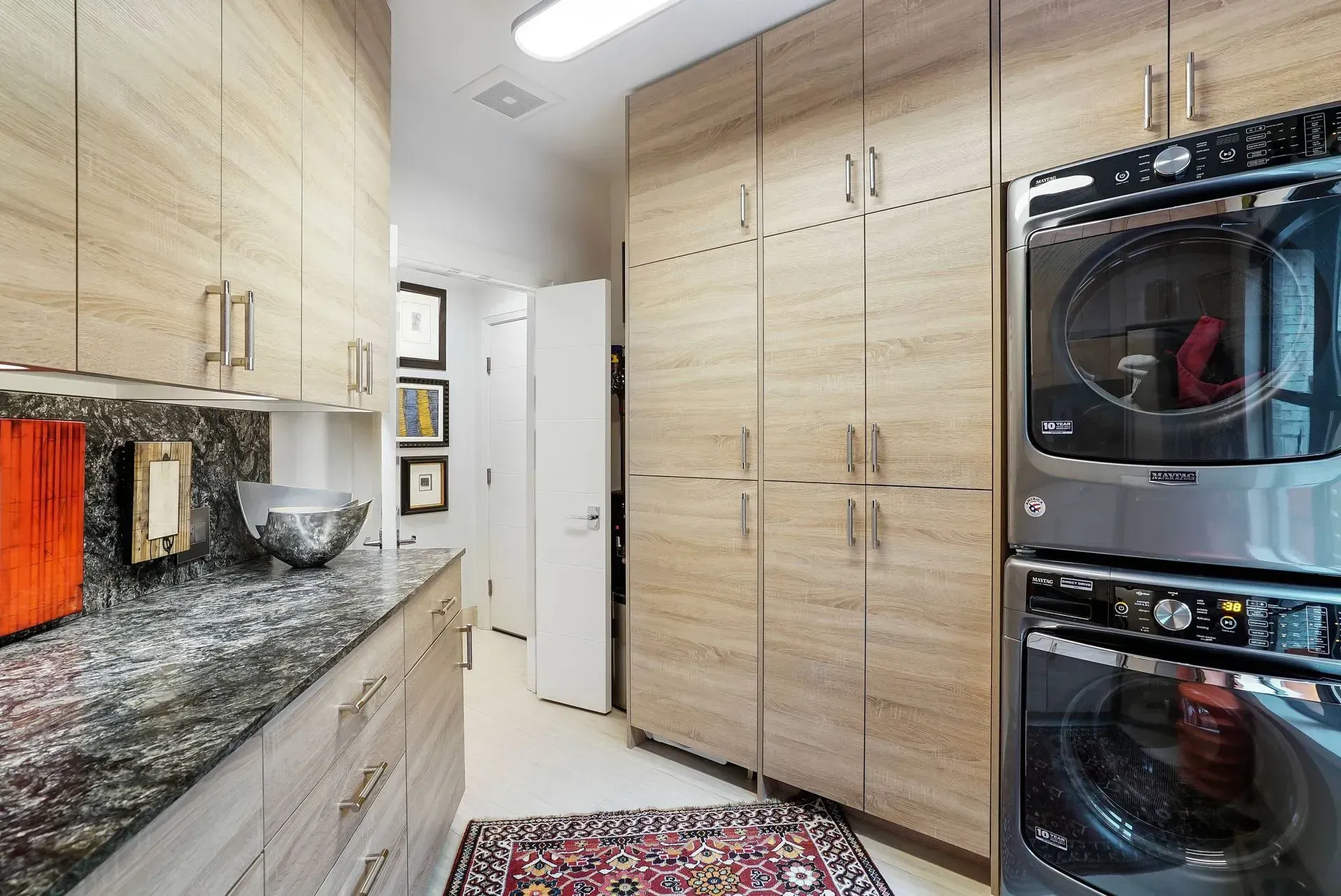 Bright kitchen with light wood cabinets, granite counters, white door, and stacked washer-dryer units.