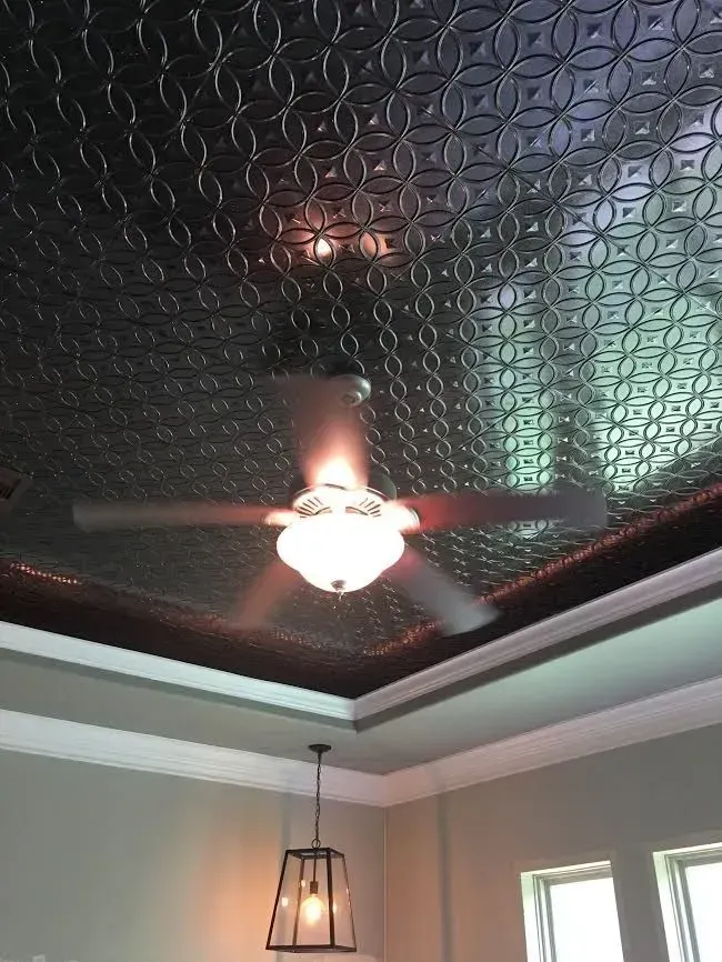 Ceiling fan and hanging light in a room with a dark patterned tray ceiling and windows.