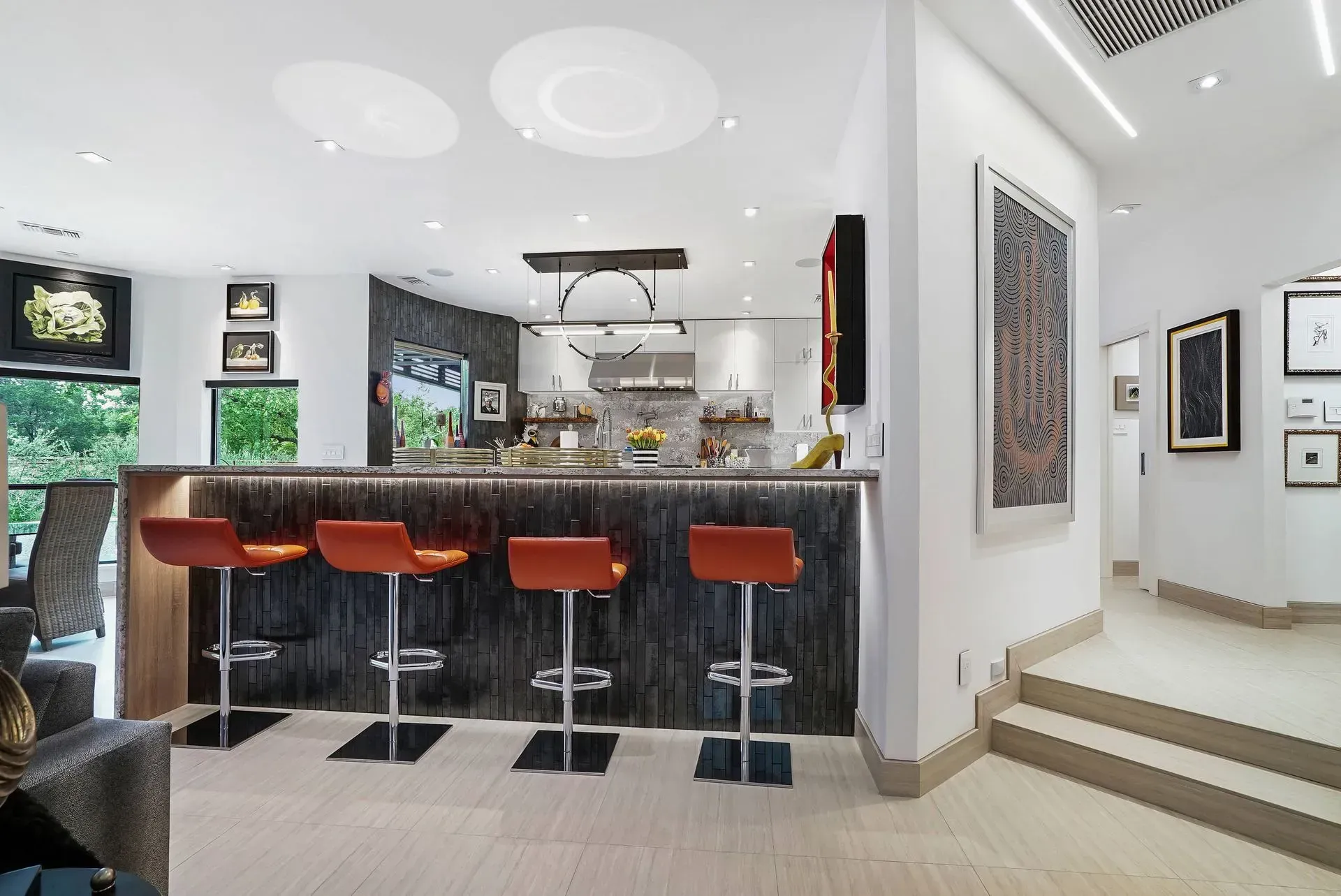 Modern kitchen with granite island, orange bar stools, and white walls leading to steps.