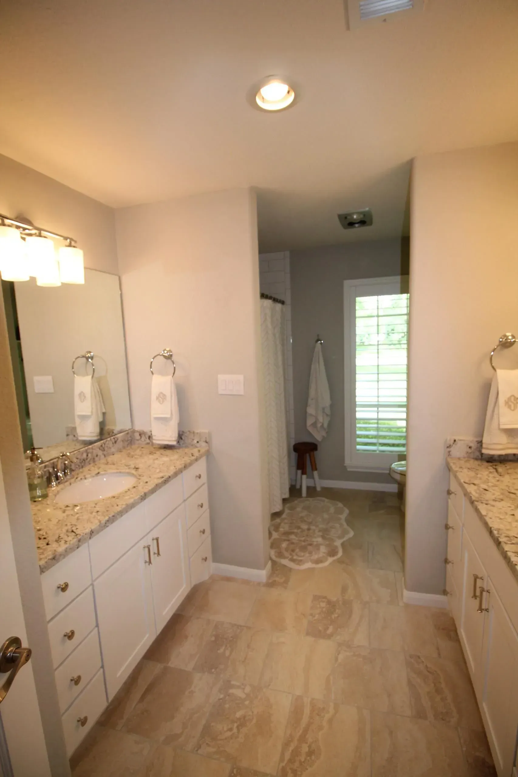 Bright bathroom with double sinks, granite counters, a central rug, and a hallway to a shower at the back.