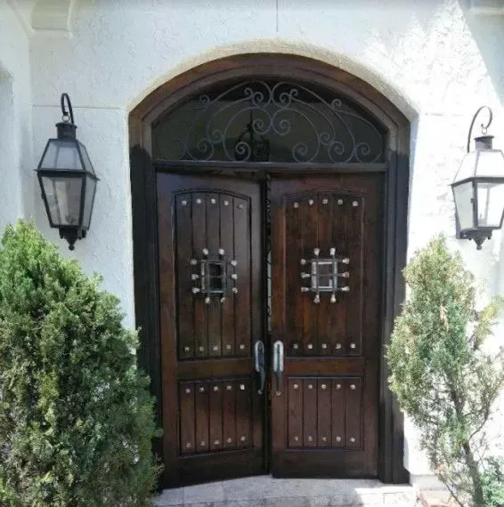Dark wooden double front door with iron details, flanked by wall lanterns and greenery.