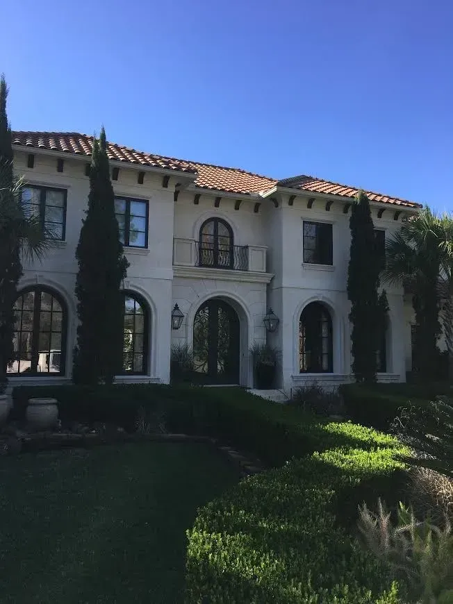 Two-story Mediterranean-style house with arched windows, red tile roof, and landscaped front yard under blue sky
