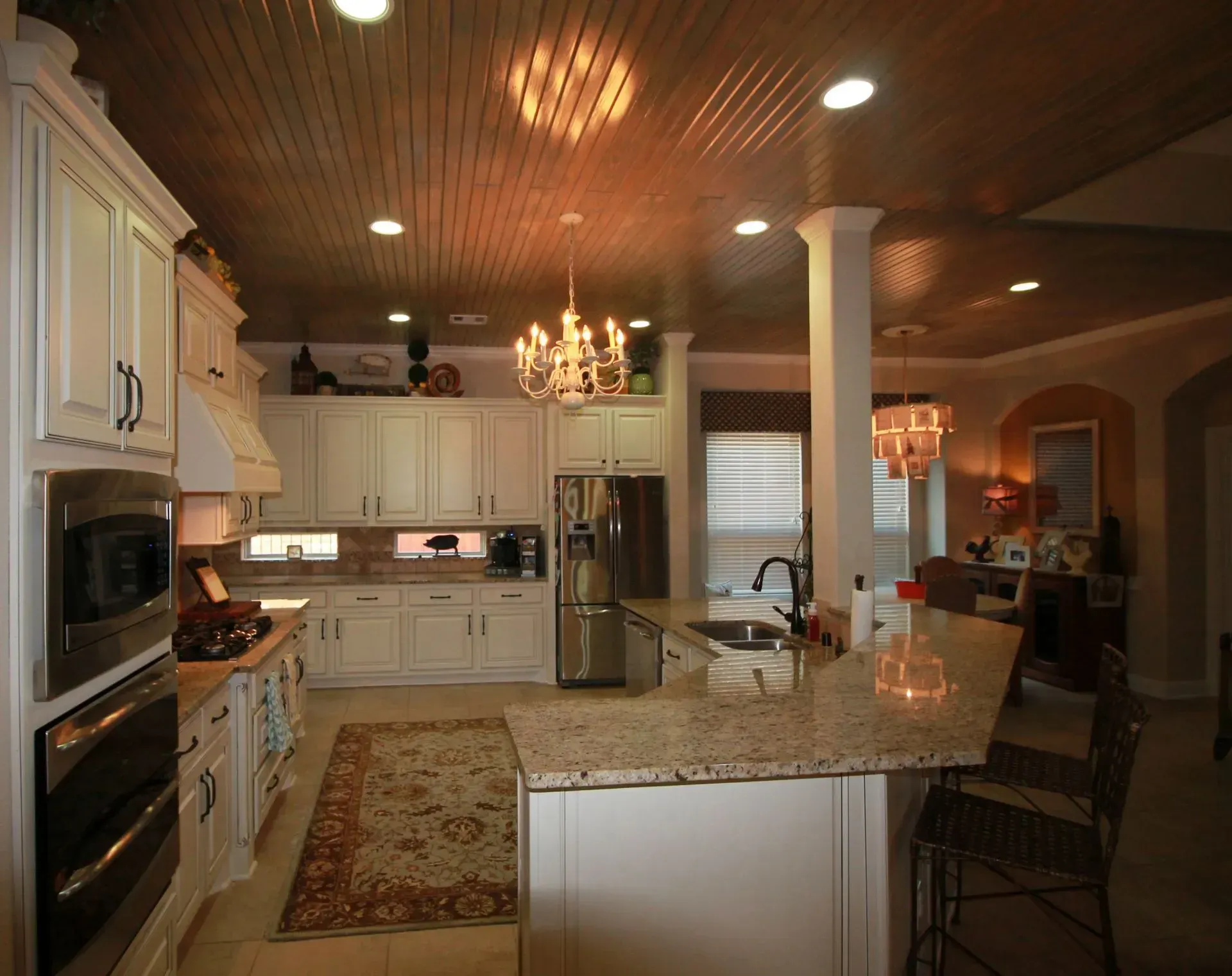Spacious modern kitchen with granite island, white cabinets, wood ceiling, and chandelier lighting.