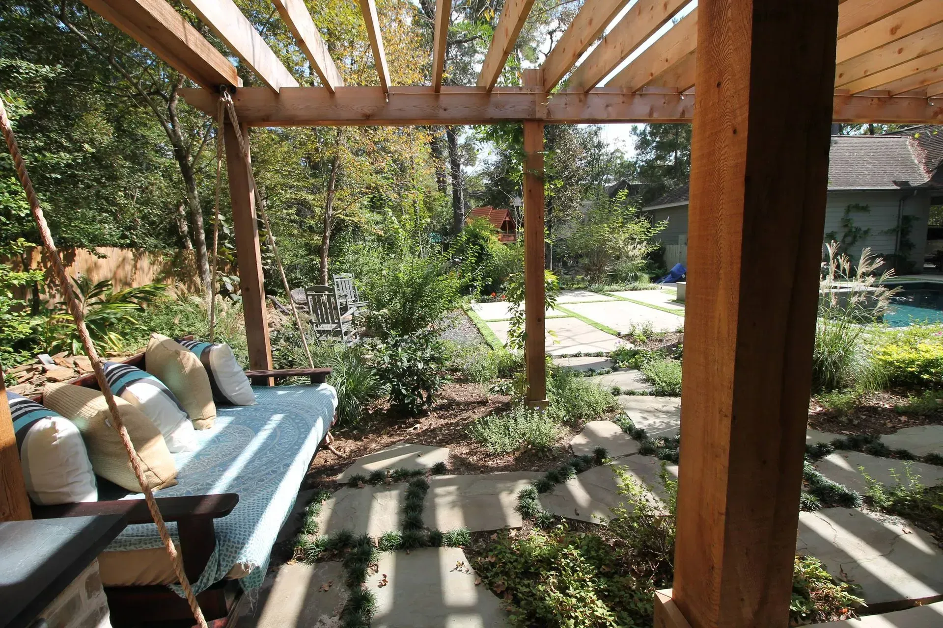 Covered patio with wooden pergola, cushioned bench, and garden path in a sunny backyard
