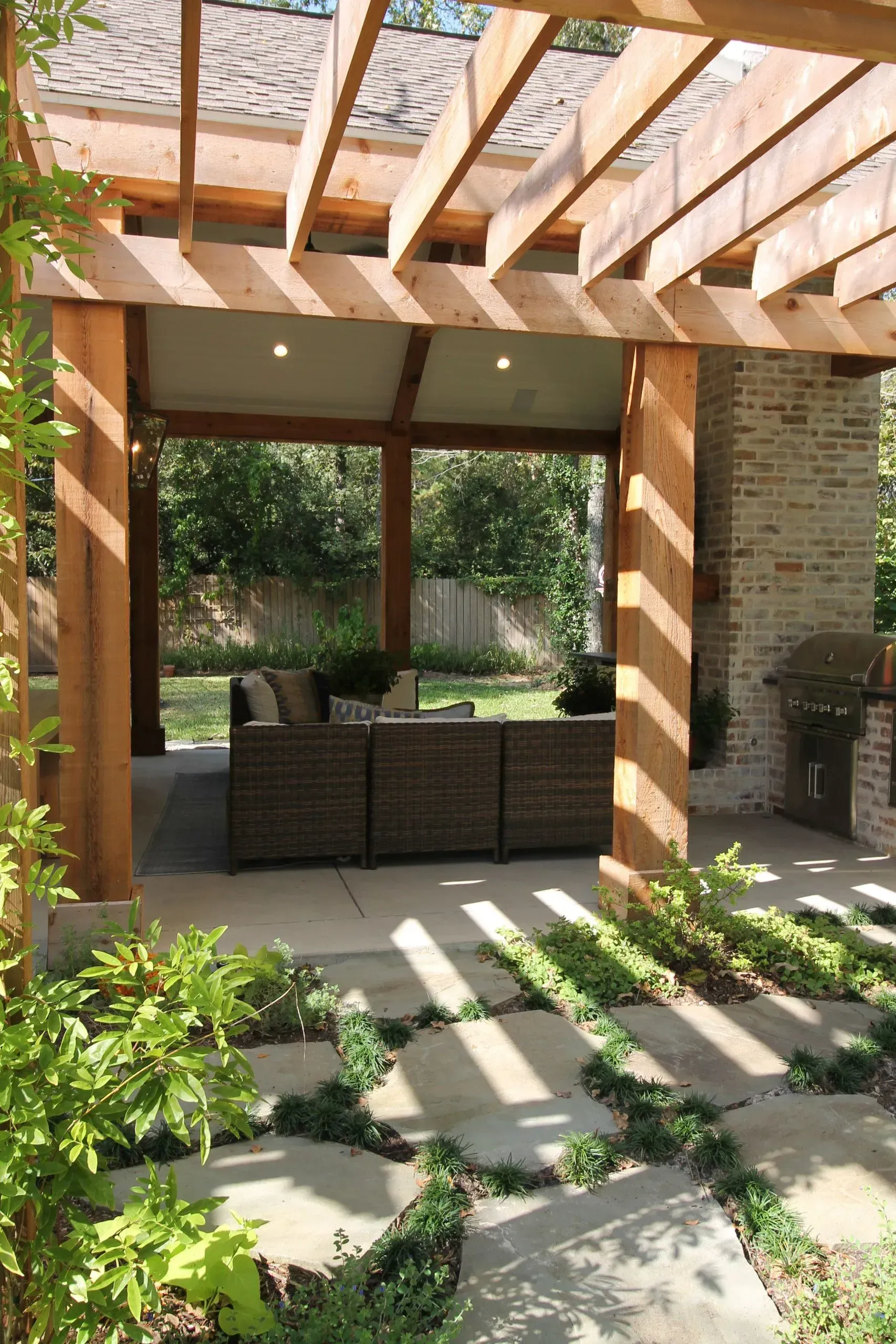 Covered patio with wicker seating, brick columns, and sunlight casting shadows across a landscaped garden.