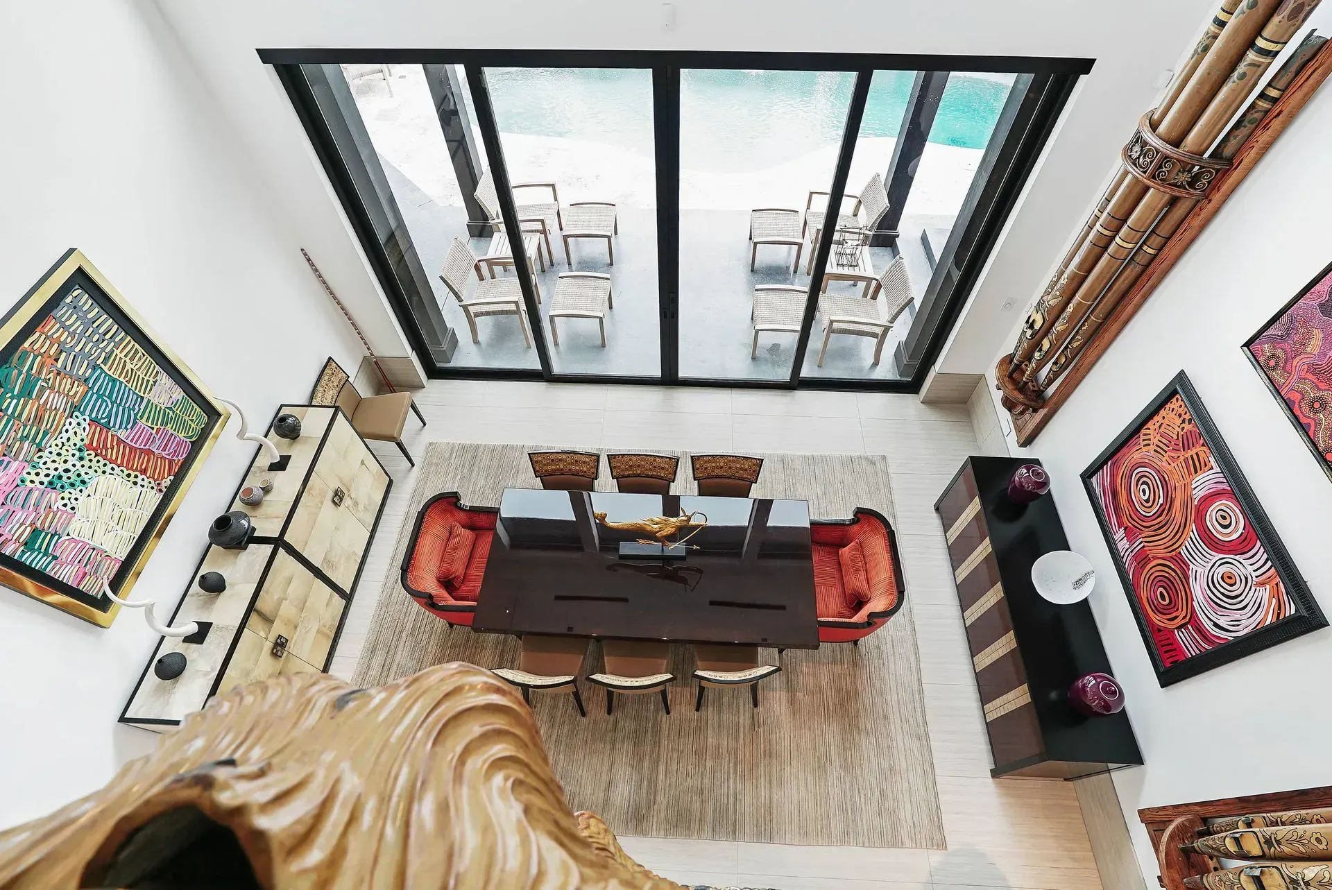 Top-down view of a modern dining room with a long table, red chairs, artwork, and large black-framed windows