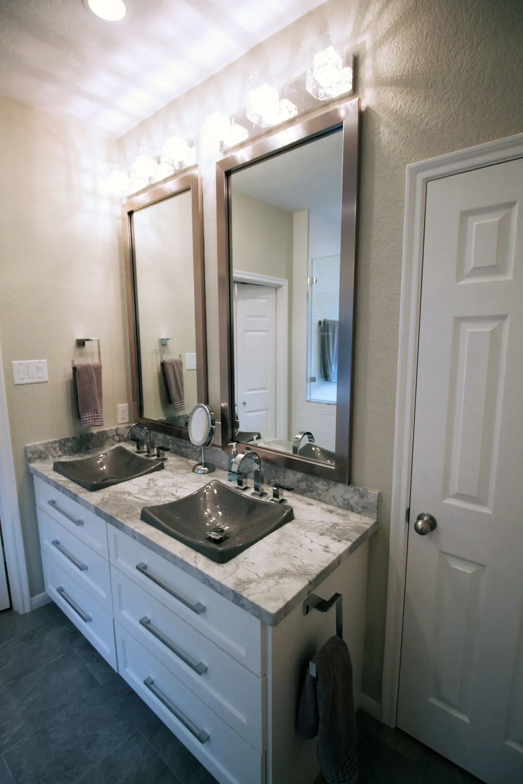 Bathroom vanity with double sinks, large mirrors, marble countertop, and wall lights beside a white door