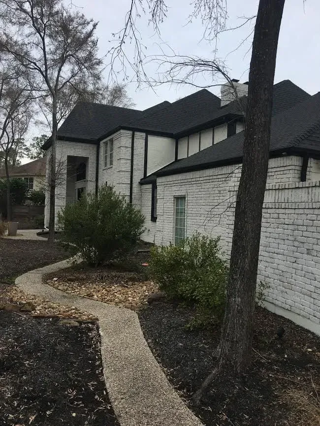 Suburban house with stone exterior, winding walkway, shrubs, and leafless trees under a gray sky