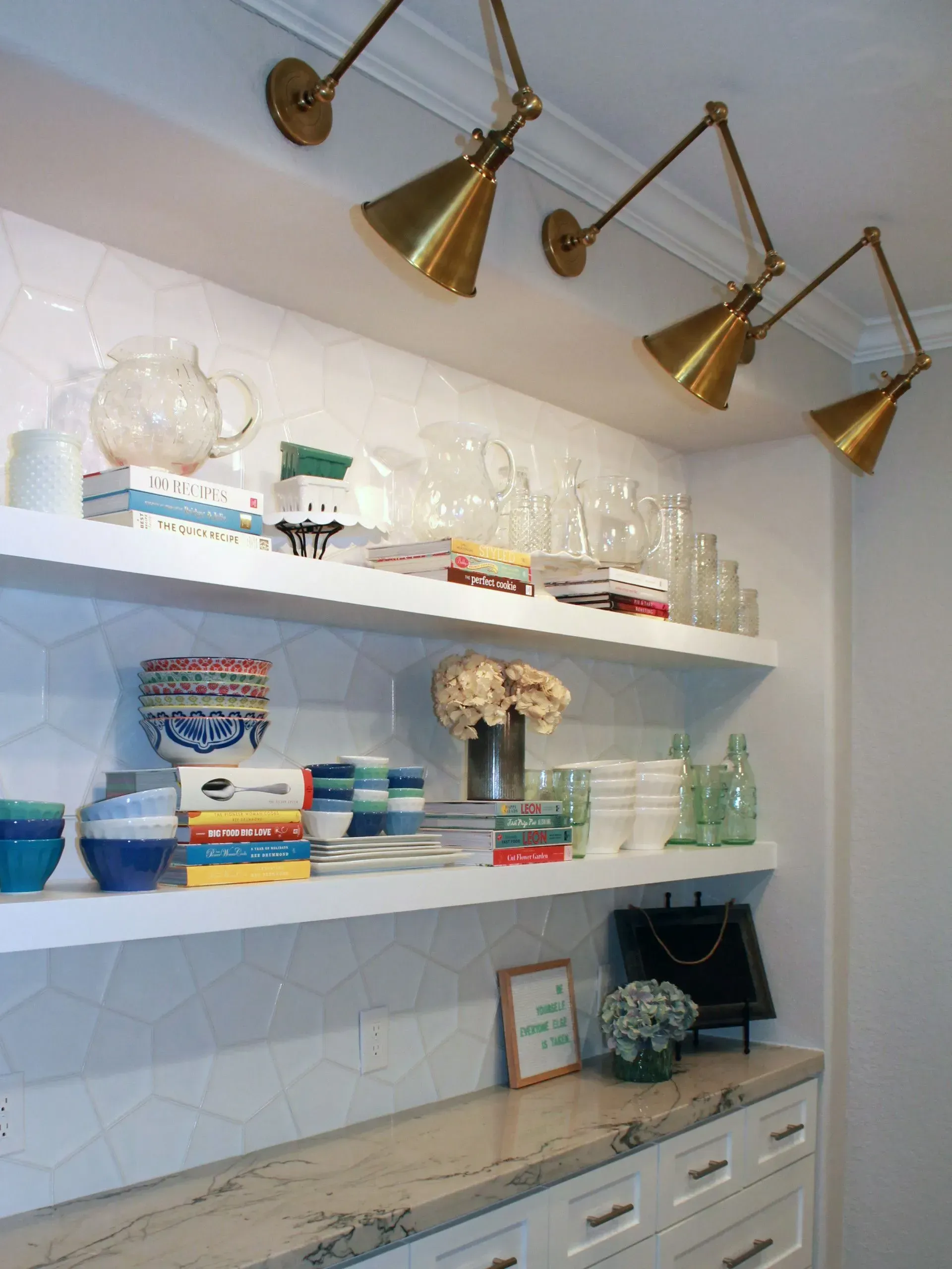 A stylish kitchen counter with open shelves displaying glassware, bowls, books, and decor under brass pendant lights