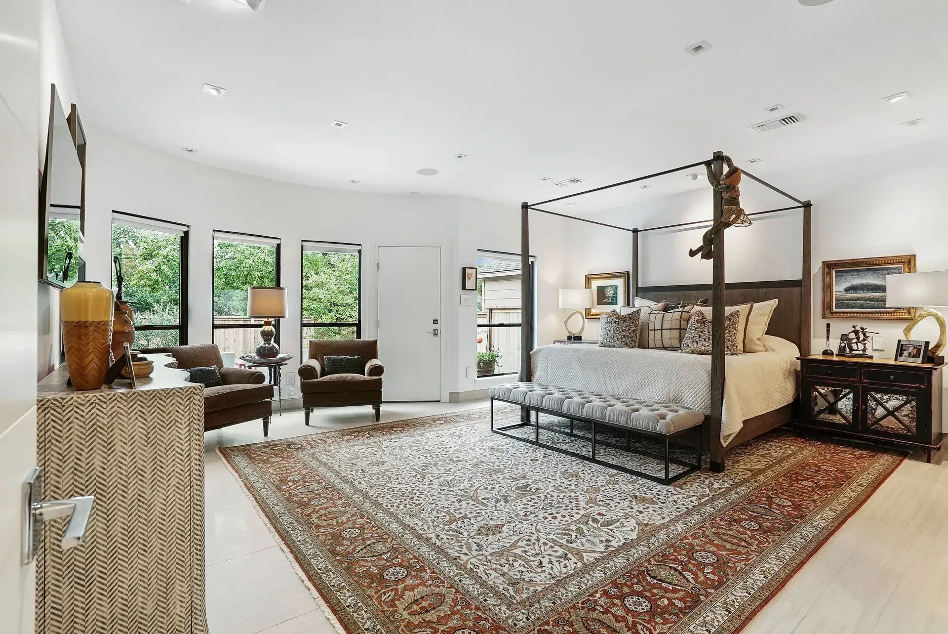 Bright modern bedroom with a four-poster bed, patterned rug, and seating by large windows.