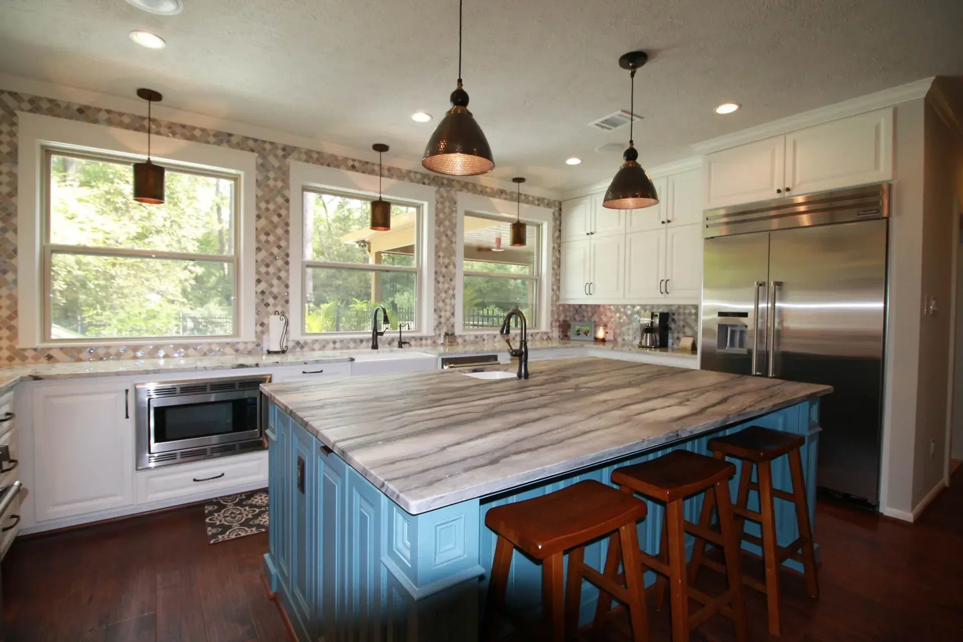 Bright kitchen with blue island, wood stools, pendant lights, and stainless steel fridge by large windows