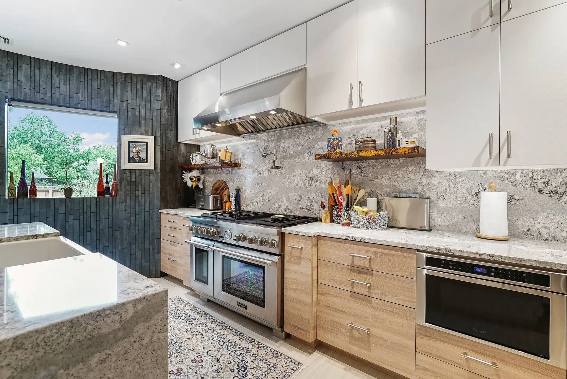 Modern kitchen with light wood cabinets, granite counters, stainless steel appliances, and a window view of greenery