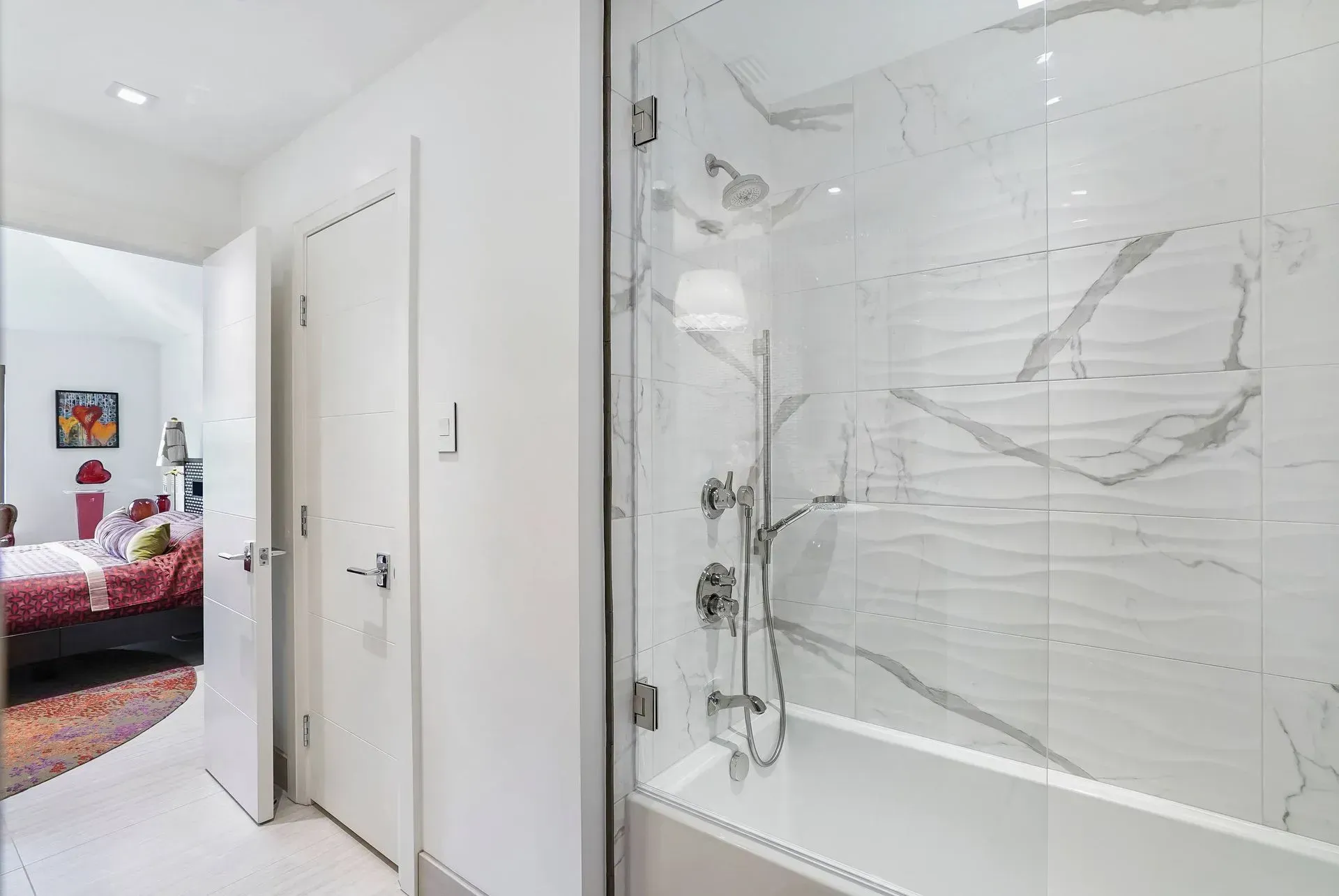 Bright hallway bathroom with white vanity, glass shower, and an open door to a bedroom with pink bedding.