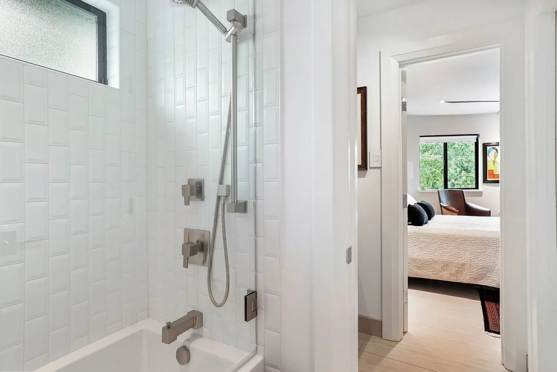 Bathroom with white tiled shower and an open door to a bright bedroom with a bed and window.