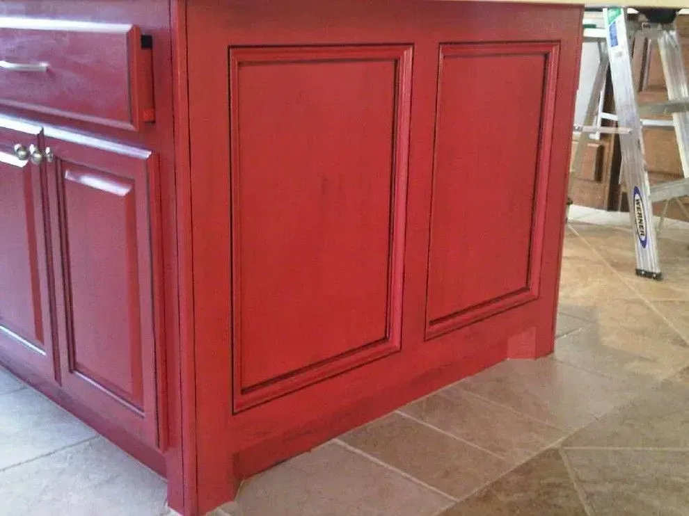 Red kitchen island cabinet with decorative paneling on a tiled floor, near a ladder in the background