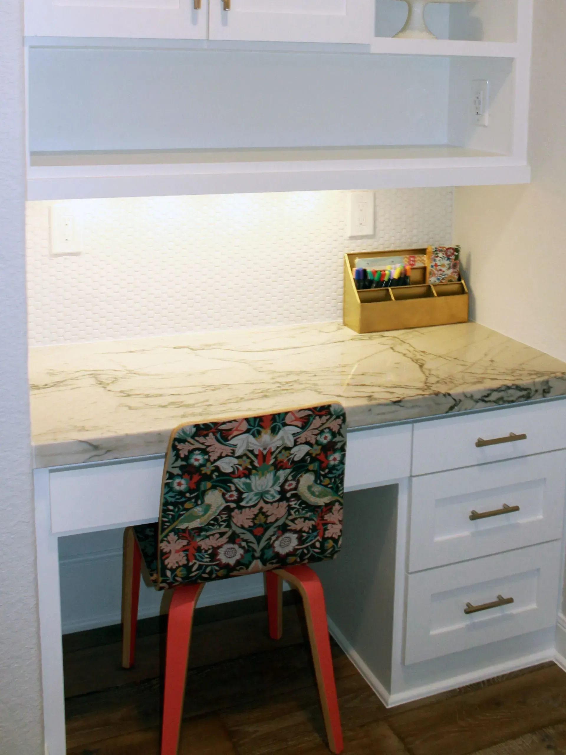 White desk with marble top, floral chair, drawers, and a shelf in a bright home office.