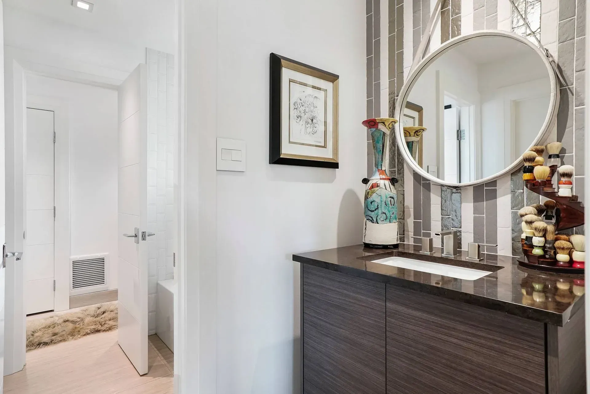 Bright hallway with a dark dresser, round mirror, and framed art leading to a white room.