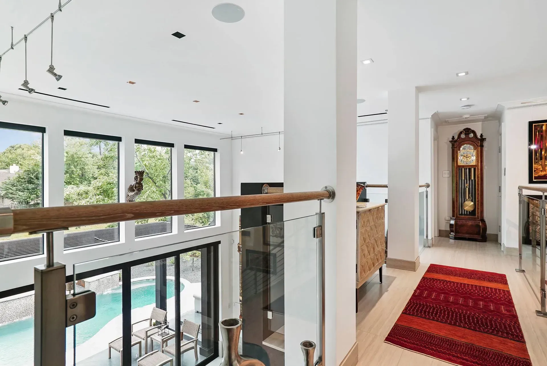 Bright modern hallway overlooking a pool, with glass railing, red runner, and a grandfather clock ahead.