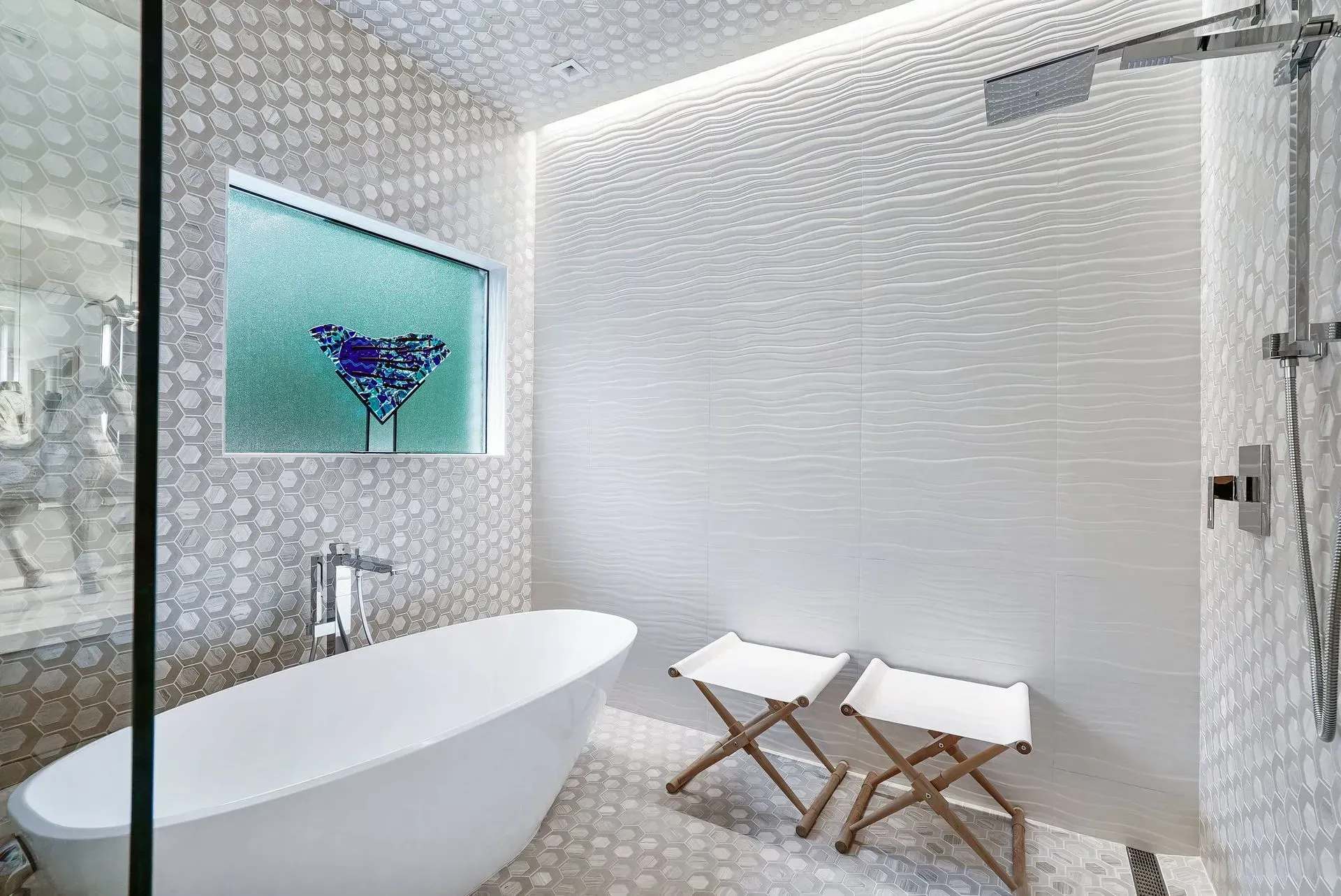 Modern white bathroom with freestanding tub, glass shower, mosaic tile walls, and two stools.