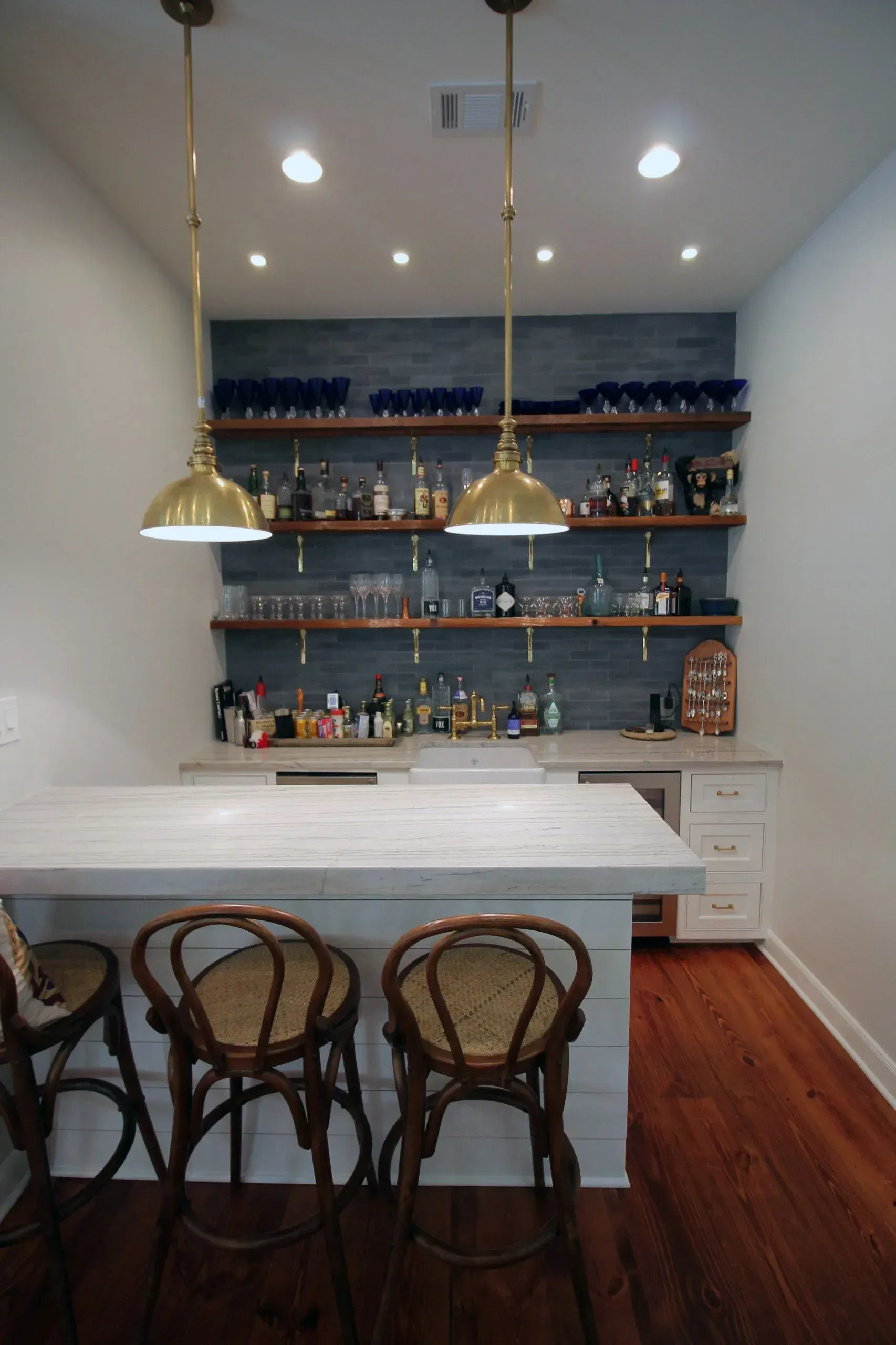 Small home bar with white counter, wicker stools, brass pendants, and blue shelves of bottles and glasses.