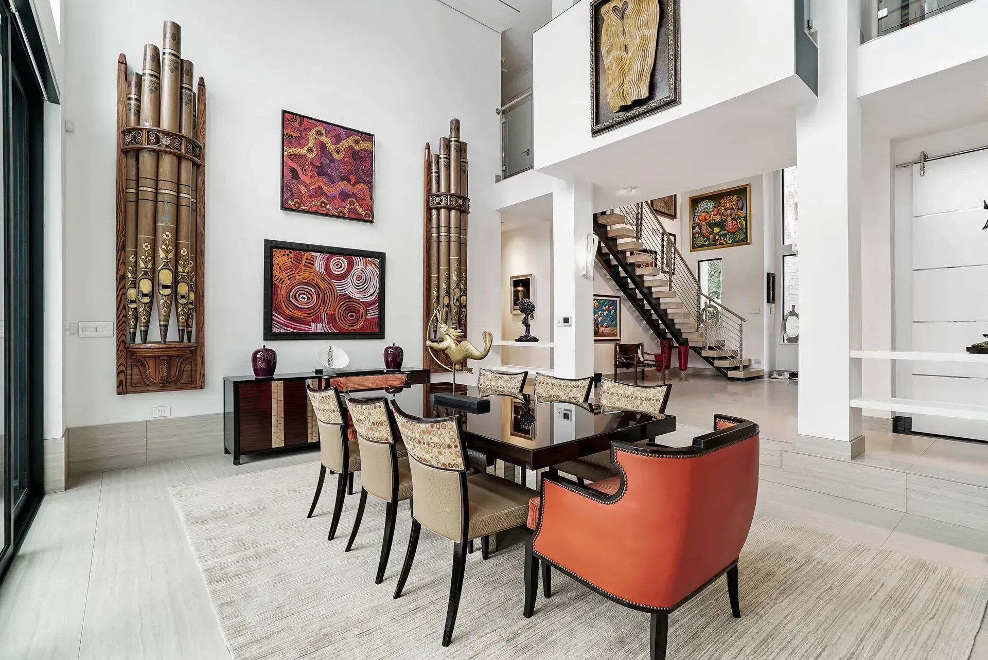 Modern dining room with high ceilings, wall art, staircase, and orange accent chairs around a long table