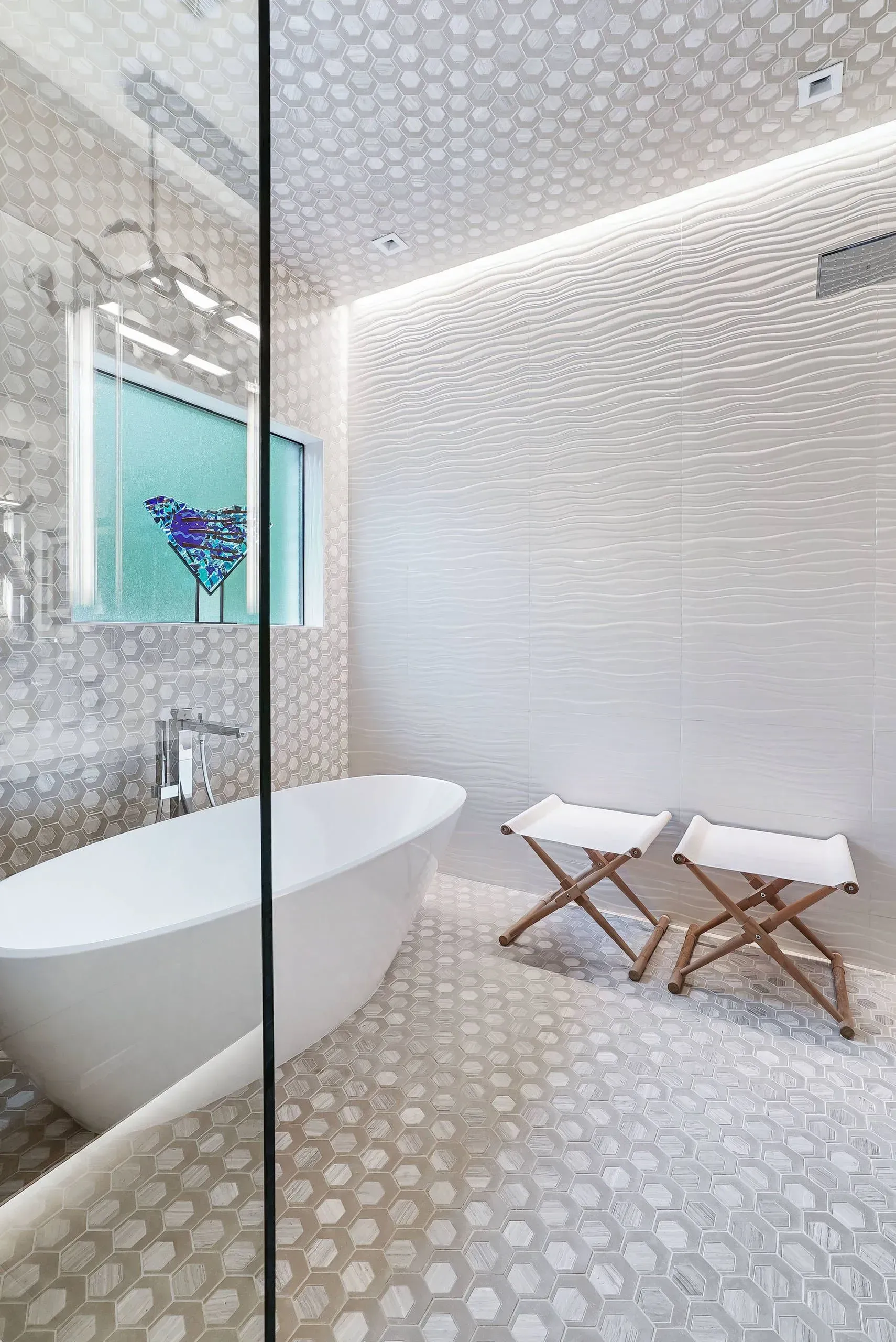 Modern white bathroom with freestanding tub, mosaic tile, and two wooden stools with folded towels