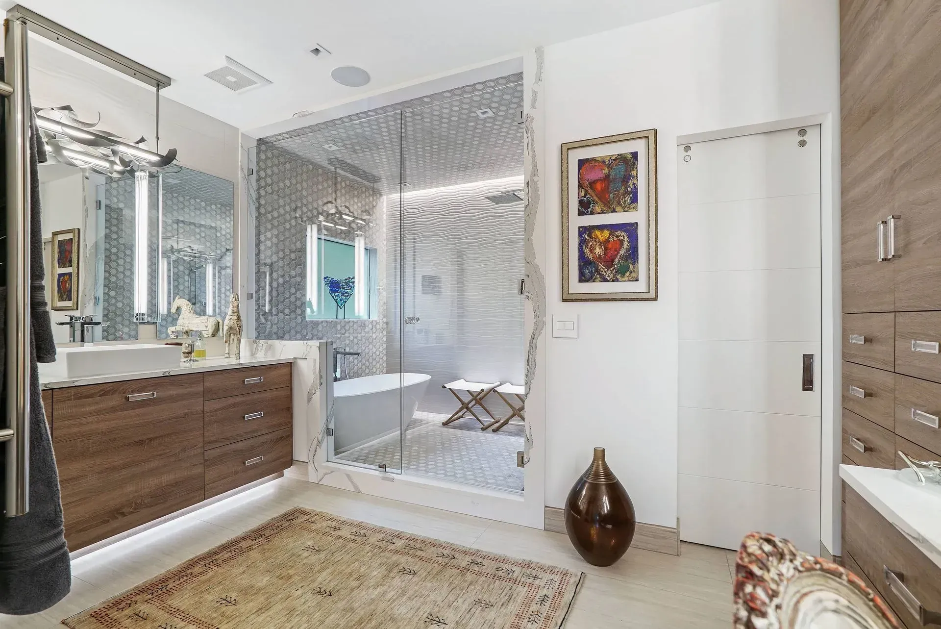 Modern bathroom with mosaic glass shower, double vanity, and white tub; framed art and patterned rug.