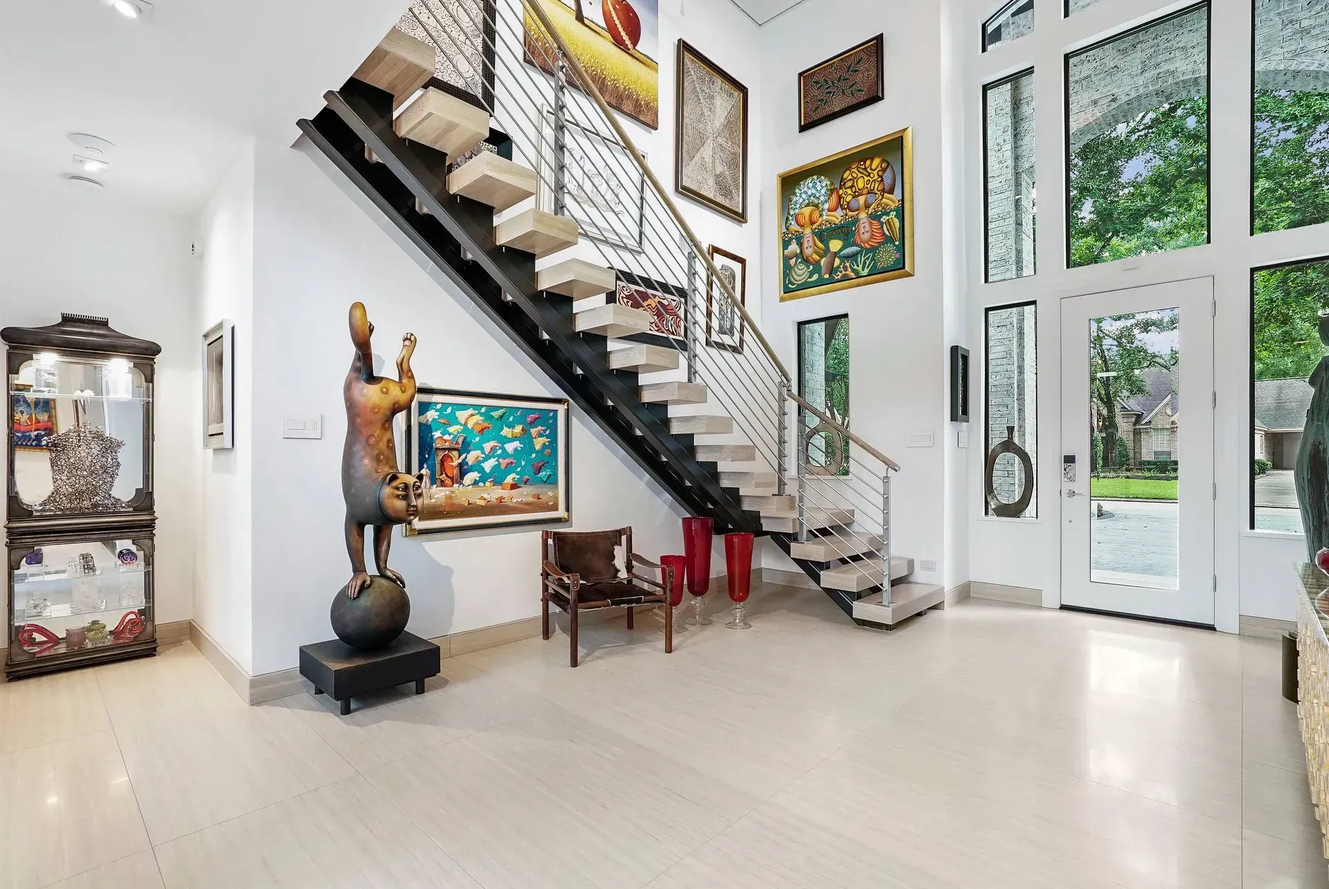 Bright modern foyer with staircase, wall art, sculpture, and glass doors overlooking greenery