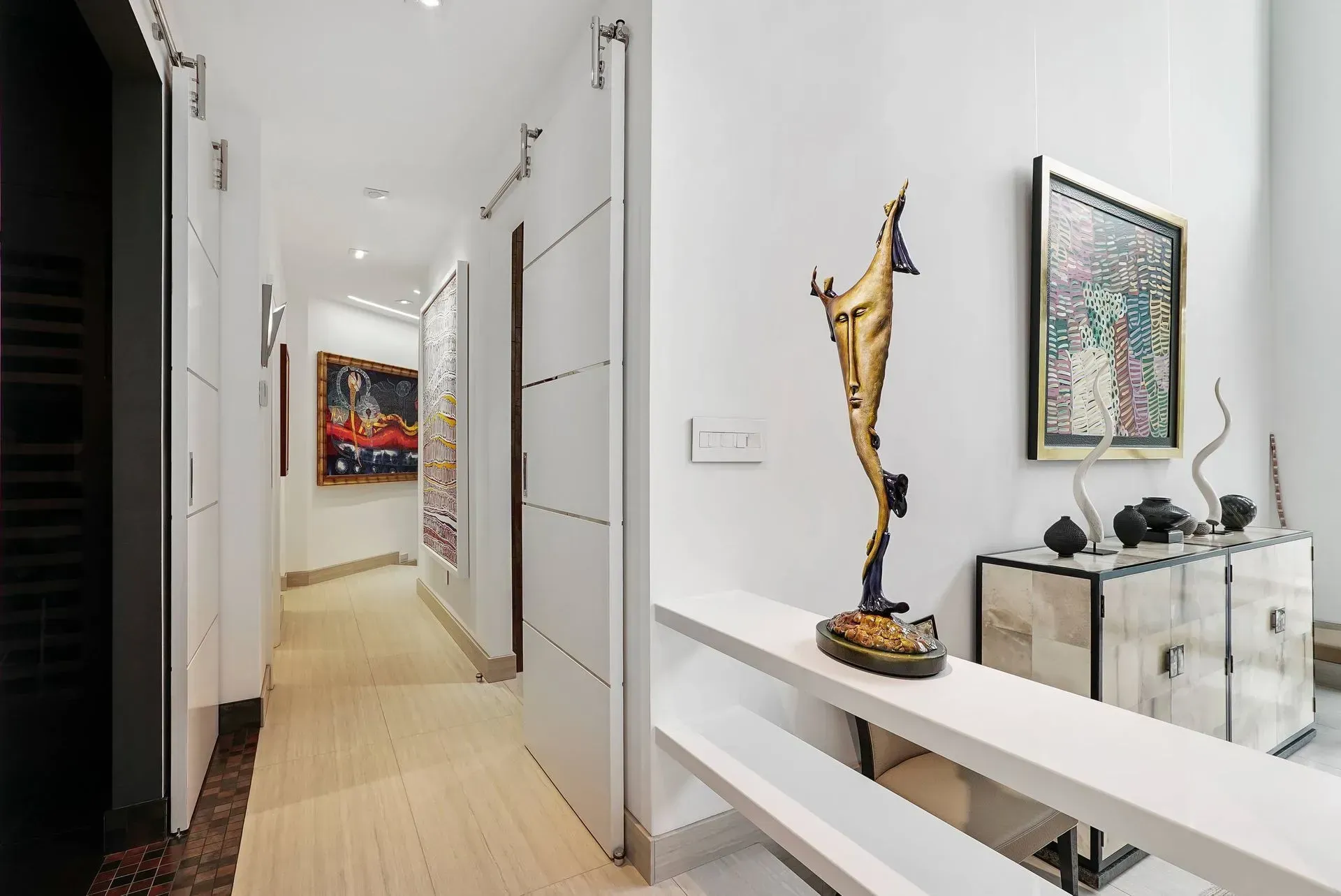 Modern hallway with white walls, light wood floor, and wall art, leading to a bright room.