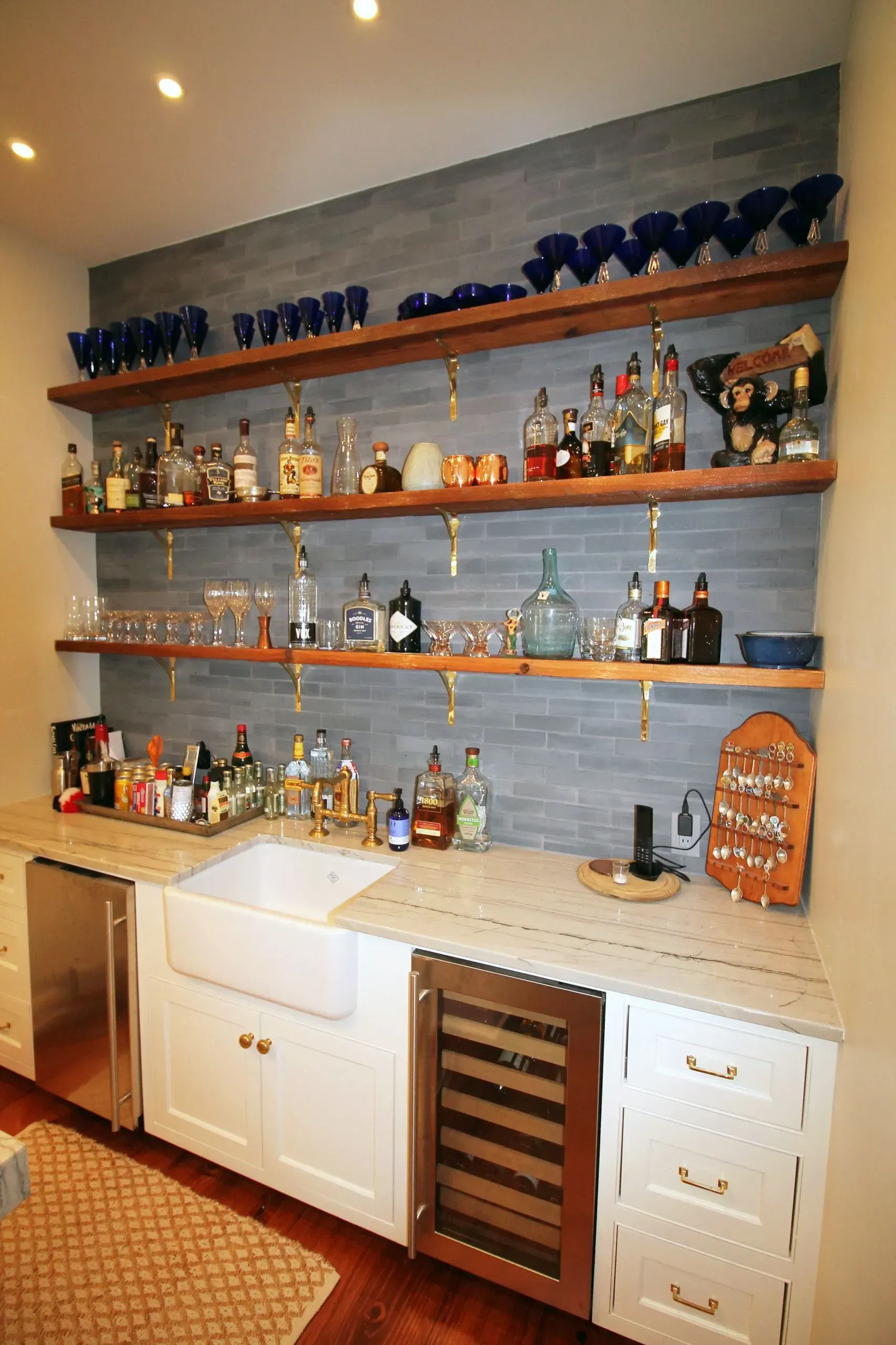 Built-in home bar with wooden shelves, glassware, liquor bottles, and white cabinets against a gray wall