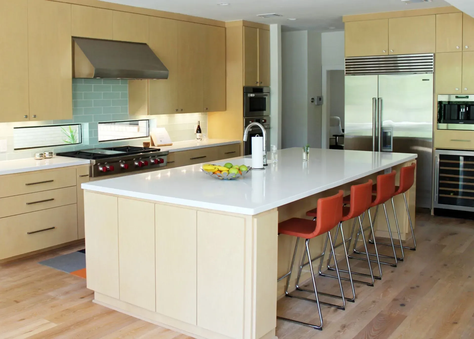 Modern kitchen with a large island, orange stools, stainless steel appliances, and light wood cabinets