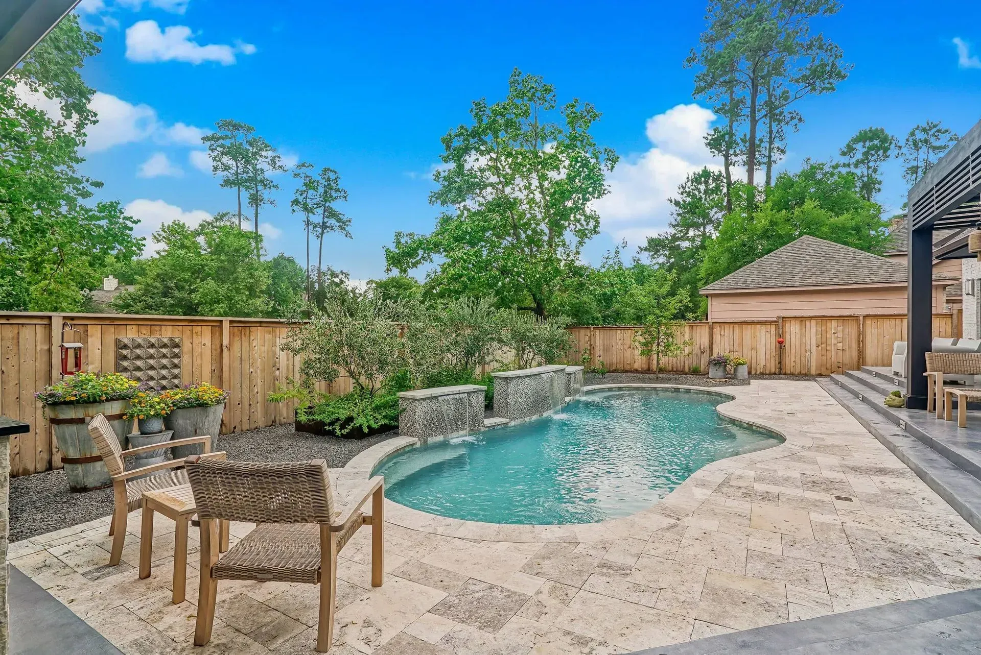 Backyard pool with stone patio, wooden fence, lounge chairs, and lush greenery under a blue sky