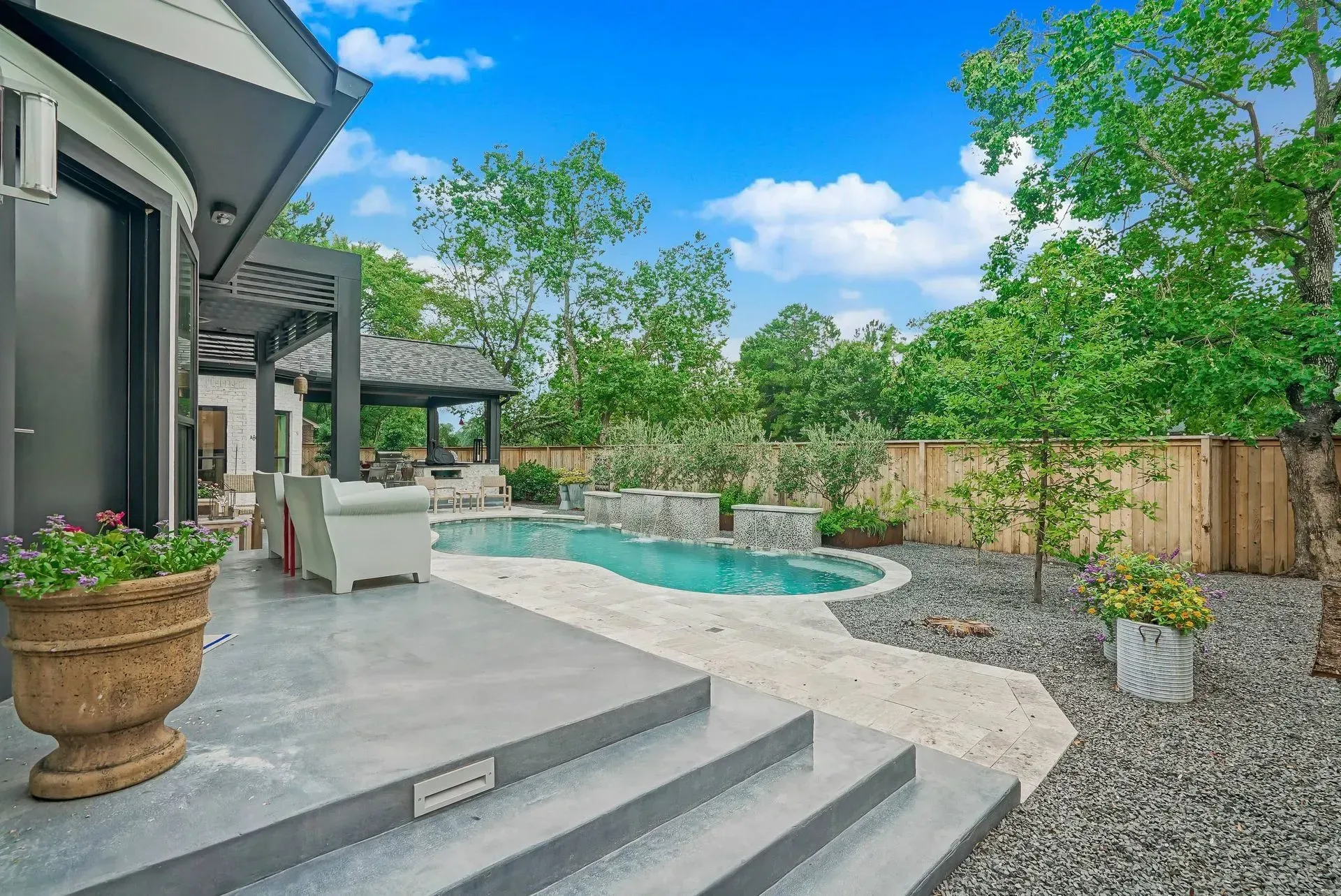 Modern backyard patio with a pool, gray steps, potted plants, and green trees under a blue sky
