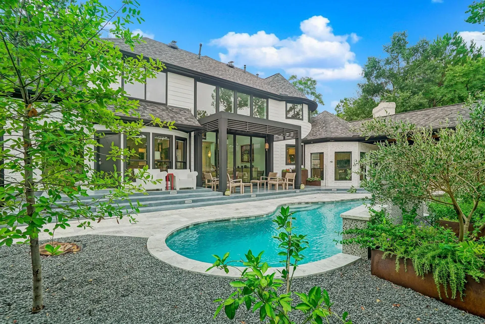 Modern house with a blue swimming pool, patio seating, and lush green landscaping under a bright sky