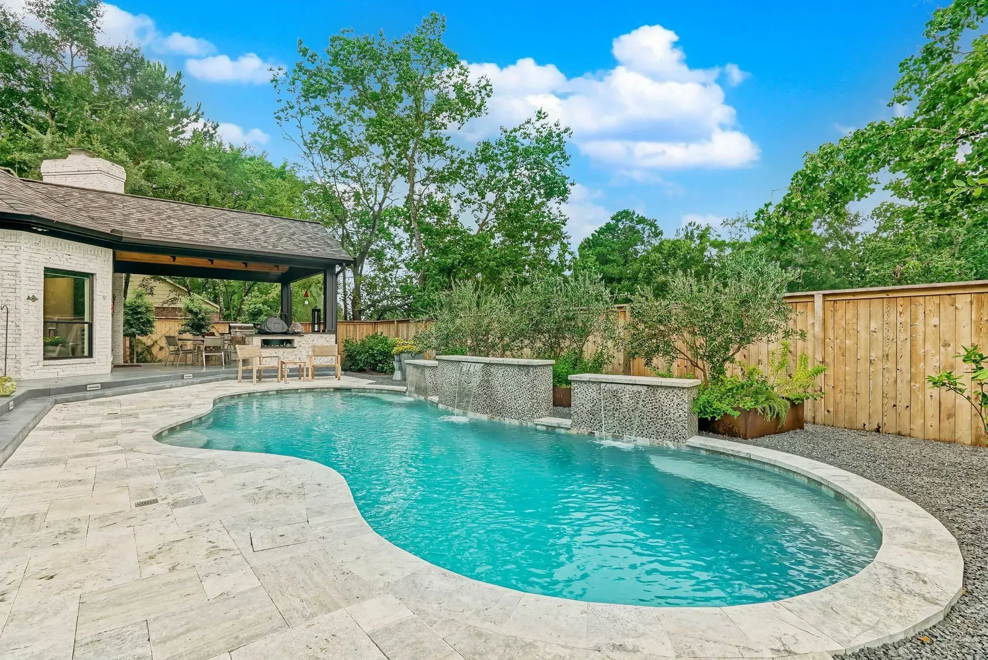 Backyard pool with stone patio, covered seating area, and trees under a blue sky