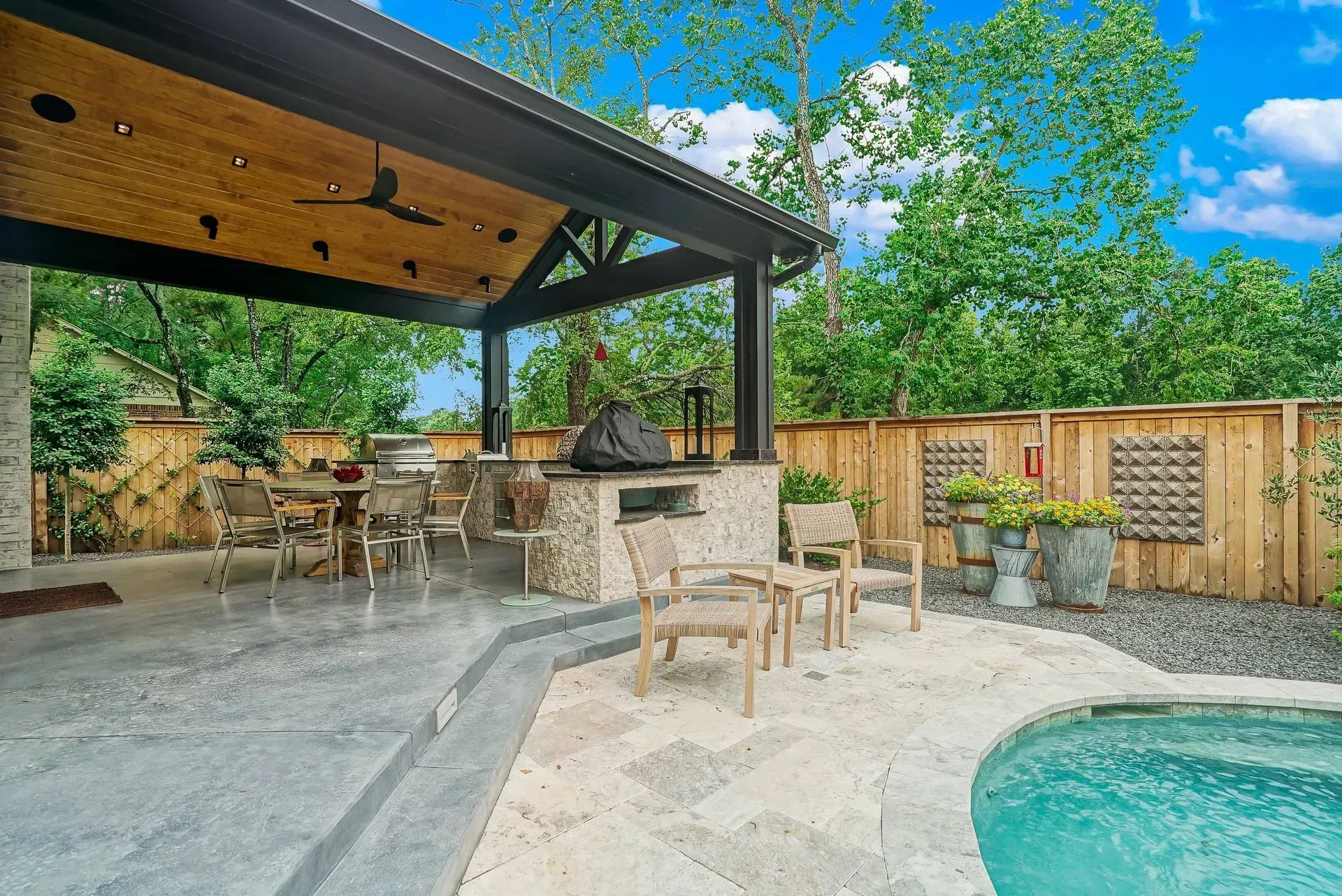 Covered patio beside a turquoise pool, with stone bar seating, wooden chairs, and a wood-beamed roof