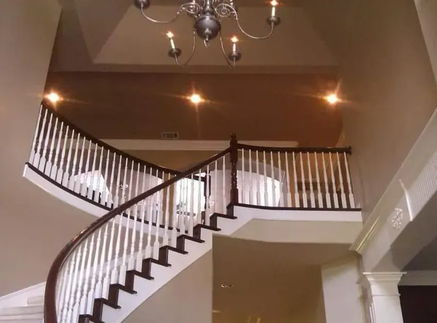 Curved staircase with white balusters and dark wood railing in a warmly lit foyer under a chandelier