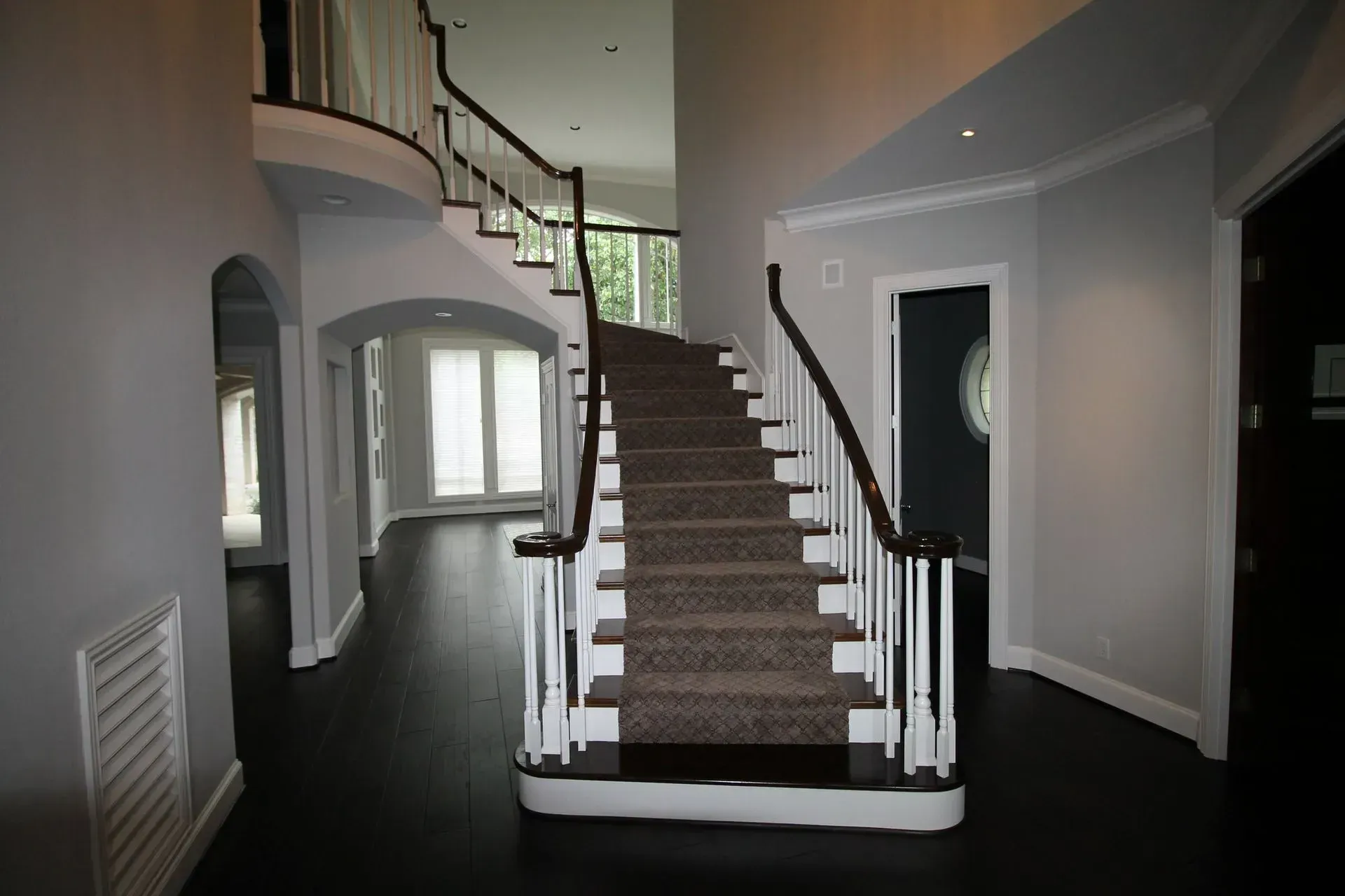 Spacious foyer with curved staircase, white walls, dark floor, and arched doorway