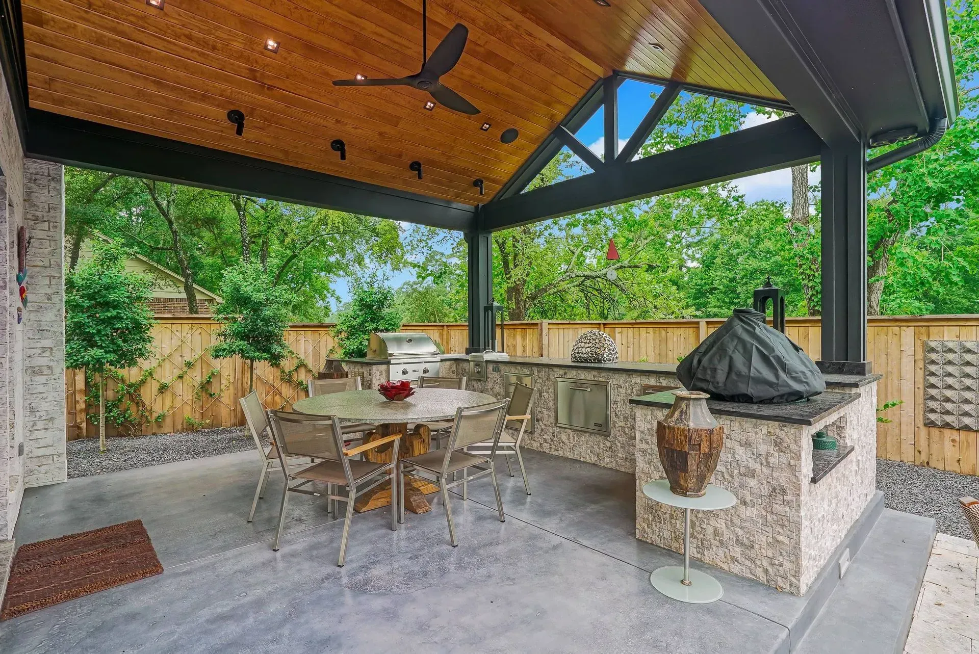 Covered outdoor patio with stone barbecue, dining table, and wooden ceiling overlooking greenery