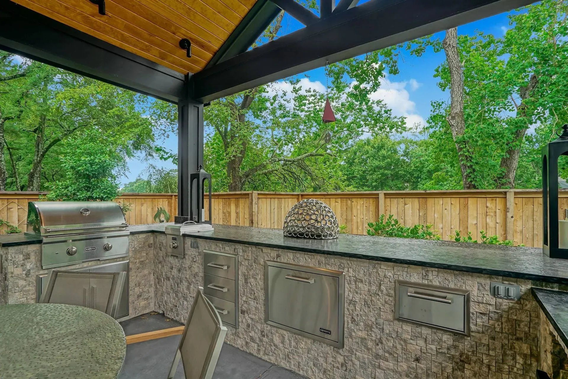 Covered outdoor kitchen with granite counters, grill, and stools under a dark roof, surrounded by trees and a wooden fence.