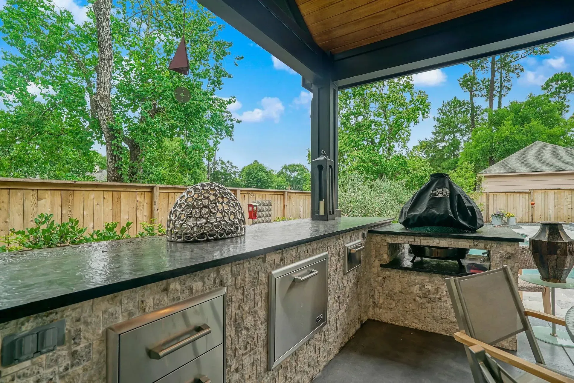 Outdoor stone bar under a covered patio with grill, stools, and backyard trees beyond the fence