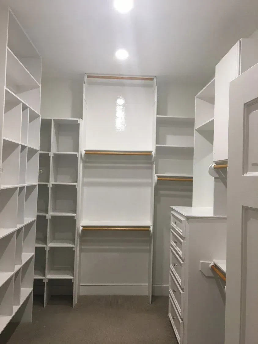 Empty white walk-in closet with shelves, hanging rods, and drawers under a ceiling light.